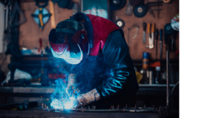 Ein Schweißer führt Schweißarbeiten an einem Metallstück auf einem Arbeitstisch durch, während Funken sprühen.