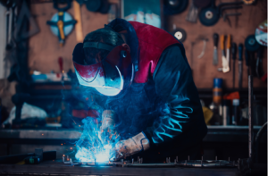 Ein Schweißer führt Schweißarbeiten an einem Metallstück auf einem Arbeitstisch durch, während Funken sprühen.
