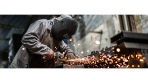 Ein Schweißer trägt Schutzbrille und Schutzhandschuhe, während er mit einer Flex an einem Metallstück arbeitet. Funken sprühen in alle Richtungen, während das Werkzeug das Metall bearbeitet. Der Schweißer ist konzentriert bei der Arbeit, umgeben von der intensiven Helligkeit und den flimmernden Funken.