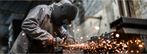 Ein Schweißer trägt Schutzbrille und Schutzhandschuhe, während er mit einer Flex an einem Metallstück arbeitet. Funken sprühen in alle Richtungen, während das Werkzeug das Metall bearbeitet. Der Schweißer ist konzentriert bei der Arbeit, umgeben von der intensiven Helligkeit und den flimmernden Funken.